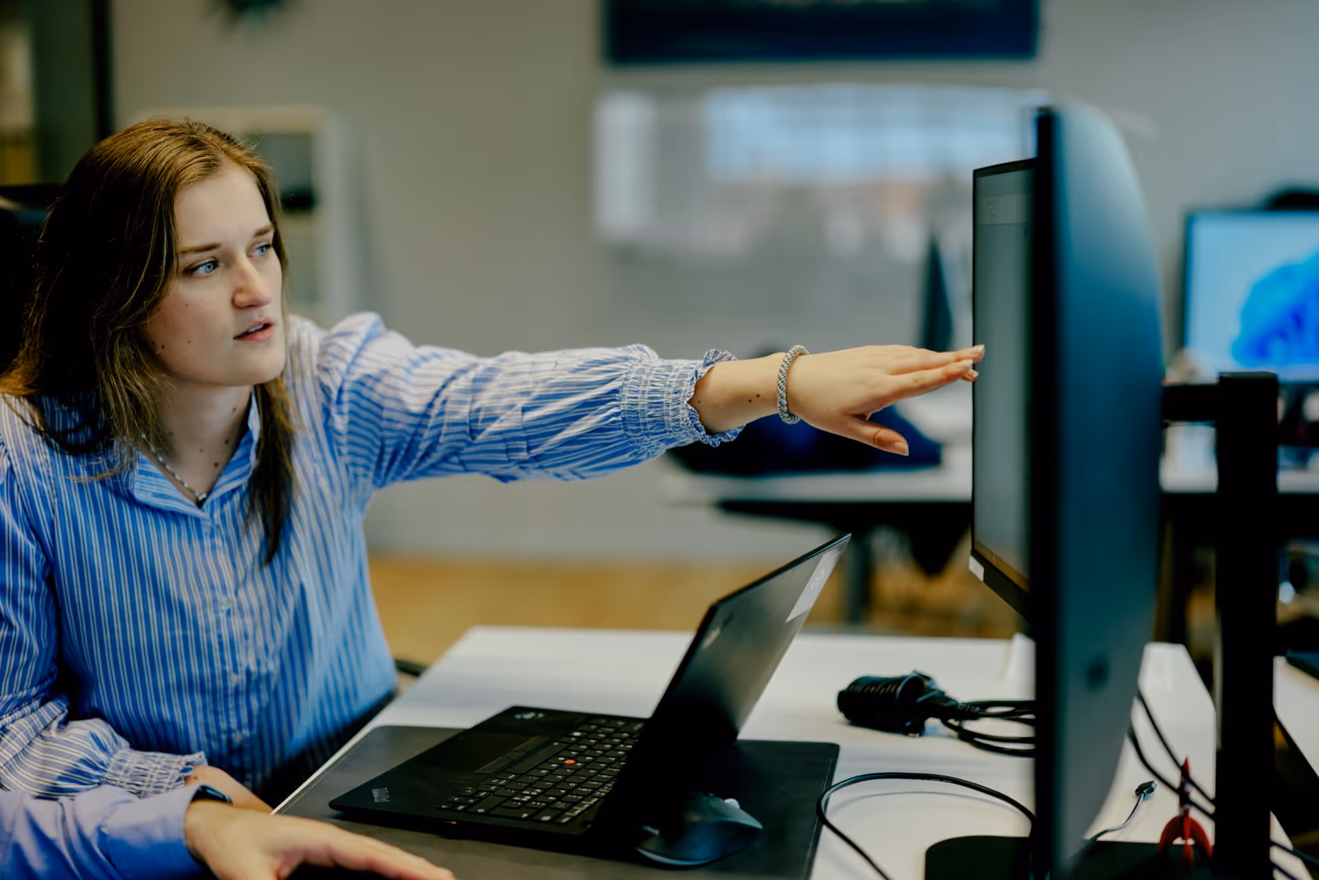 Woman in a blue striped shirt pointing at a computer monitor in an office setting.