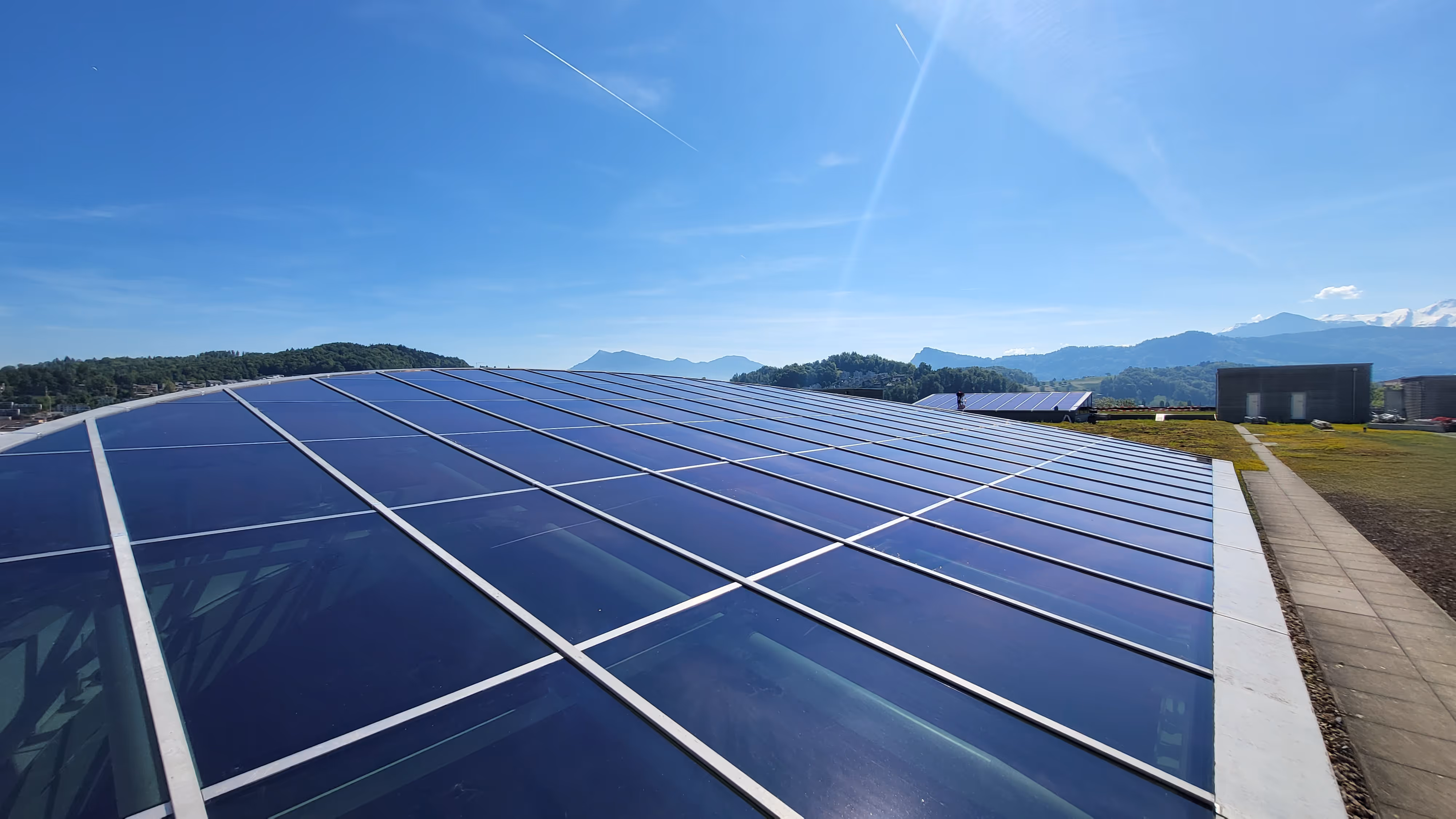 Dach mit grossen Solarpaneelen unter blauem Himmel und Bergen im Hintergrund.