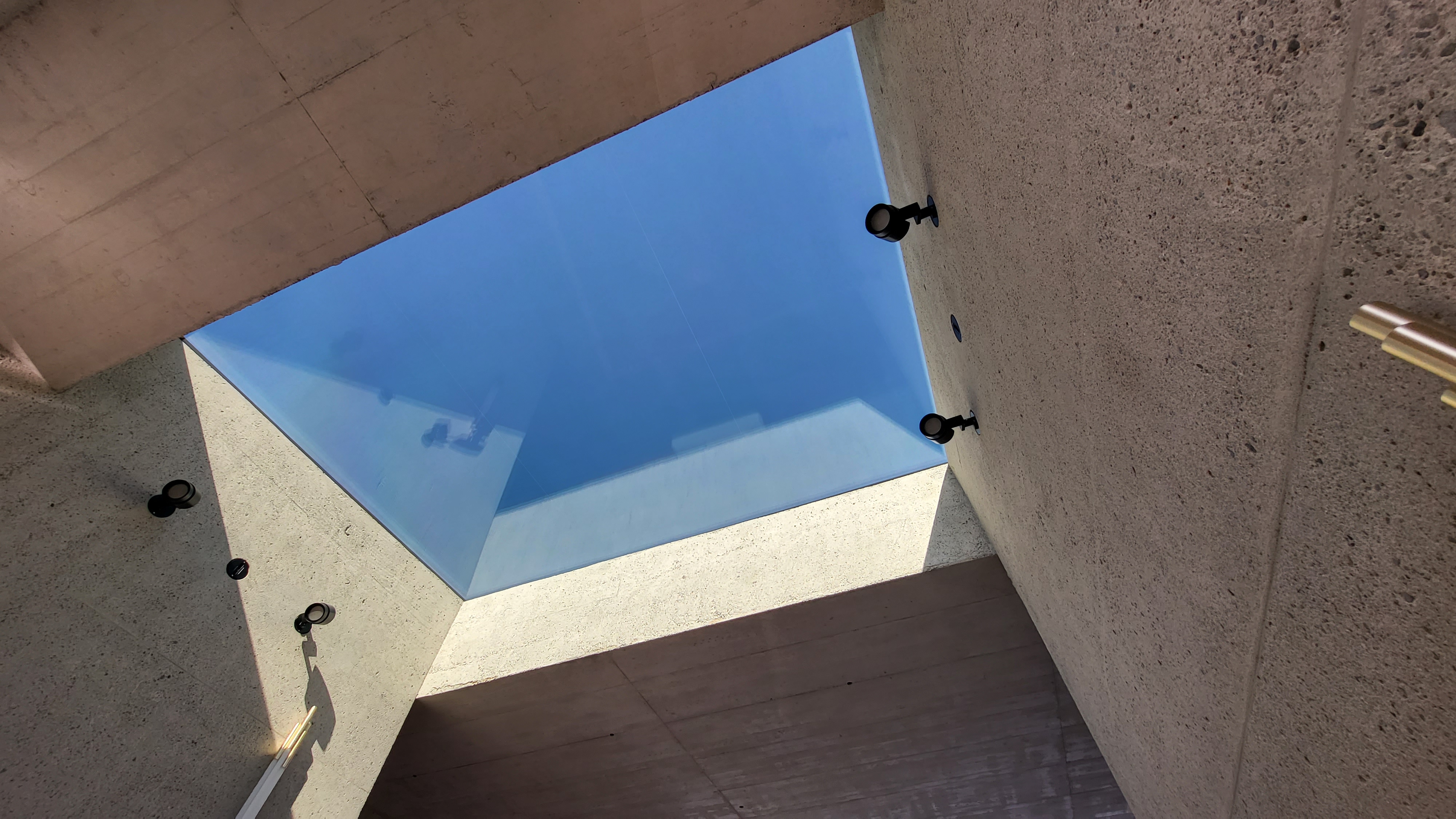 Blick nach oben durch einen quadratischen Glasdach-Ausschnitt mit Sicht auf den blauen Himmel, umgeben von Betonwänden mit Lampen.