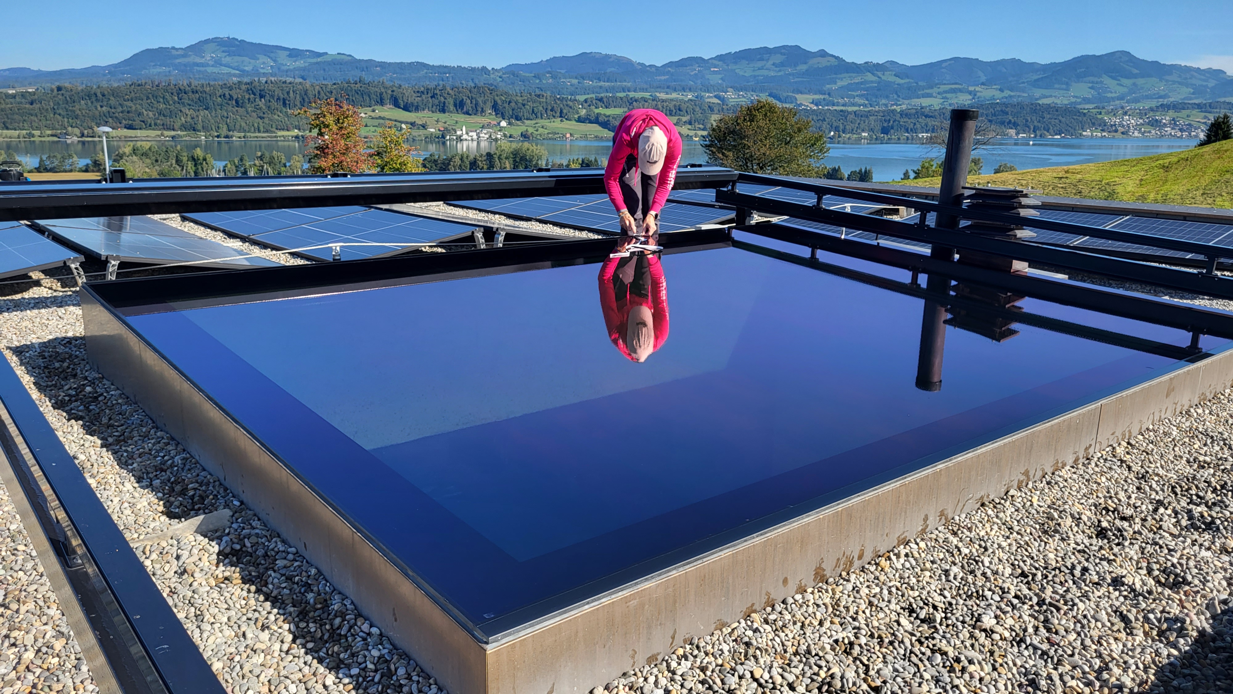 Person in pink jacket und Kappe, die auf einem flachen Solarpanel auf einem kiesbedeckten Dach arbeitet, mit Bergen und einem See im Hintergrund.