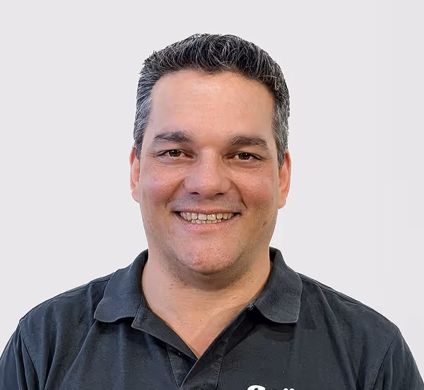 Porträt eines lächelnden Mannes mit dunklen Haaren und schwarzem Poloshirt vor hellgrauem Hintergrund.