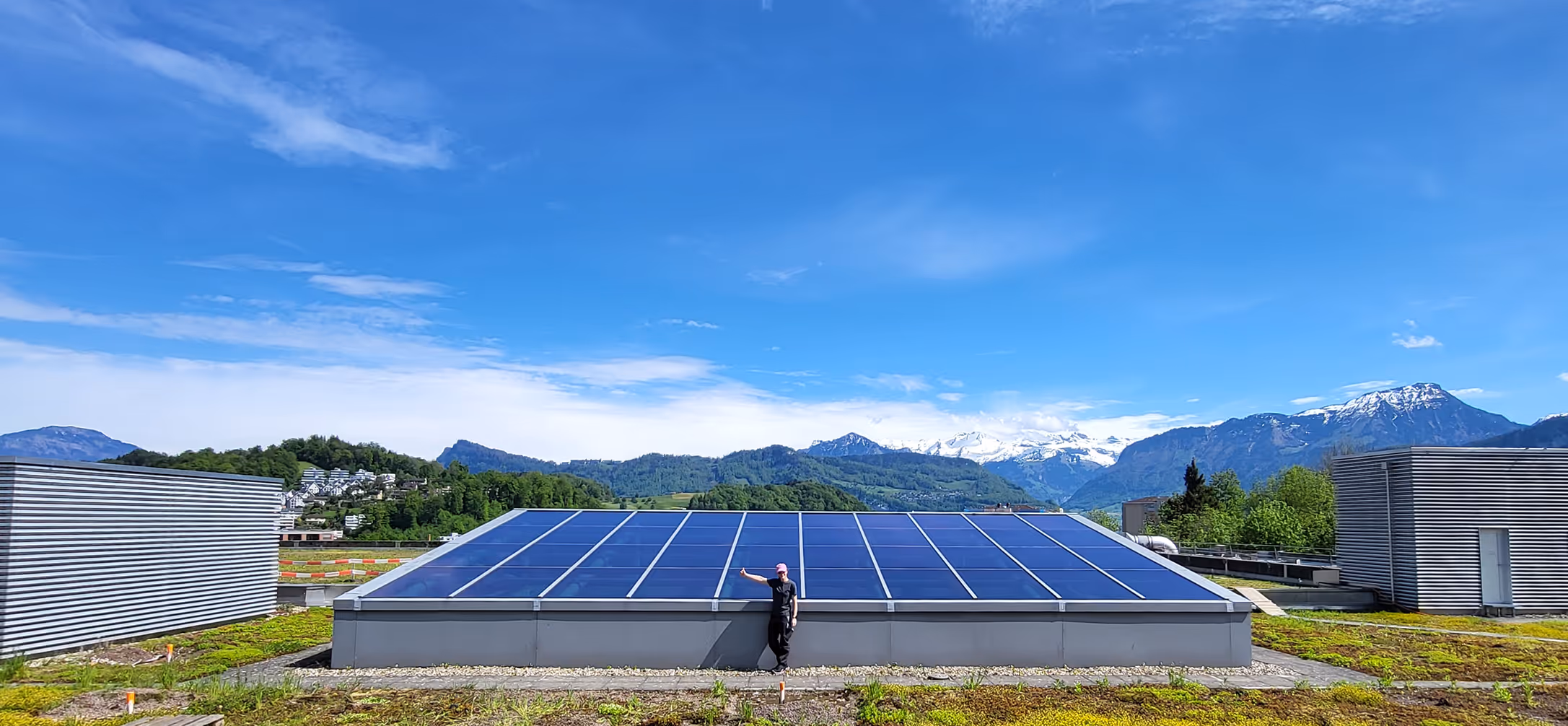 Person lehnt an grossen Solarpaneelen auf einem Dach vor Bergen mit schneebedeckten Gipfeln unter blauem Himmel.