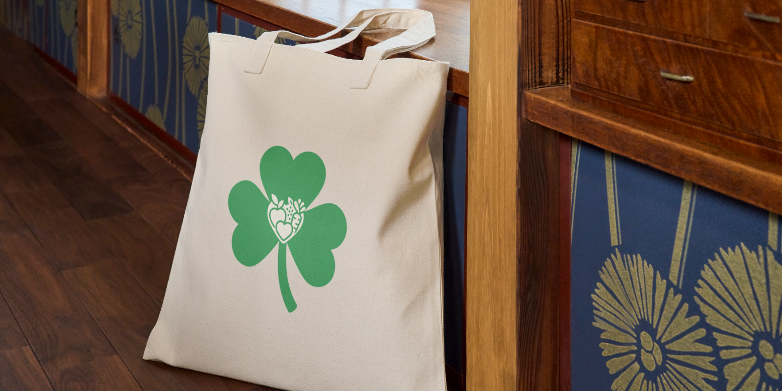 Beige tote bag with a large green shamrock and a heart-shaped fruit and vegetable design inside, resting by wood furniture.