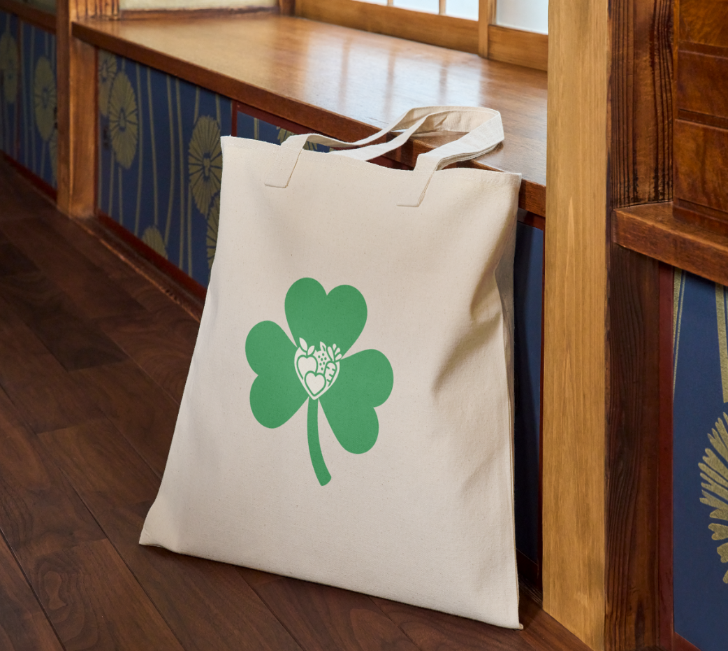 Beige tote bag with green three-leaf clover and heart-shaped icons on wooden floor near a bench.