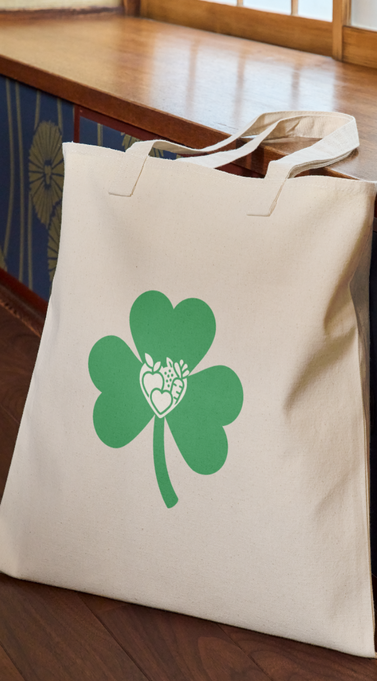 Beige tote bag with green three-leaf clover and white heart-shaped fruits and vegetables inside.
