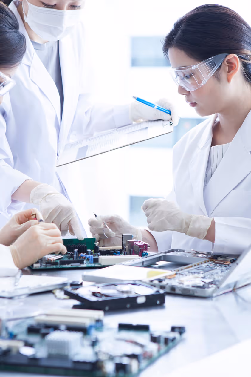 Scientists in white lab coats and safety goggles working with electronic circuit boards and taking notes.