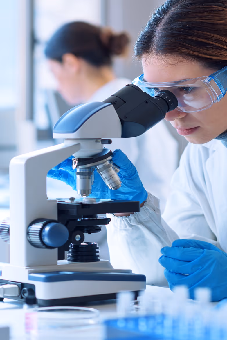 Scientist wearing safety goggles and blue gloves examining a sample under a microscope in a laboratory.