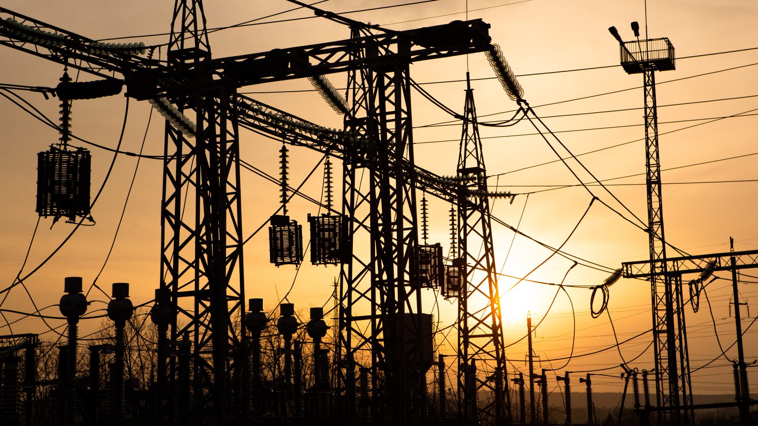 Silhouette of electrical substation equipment with power lines and transformers at sunset.