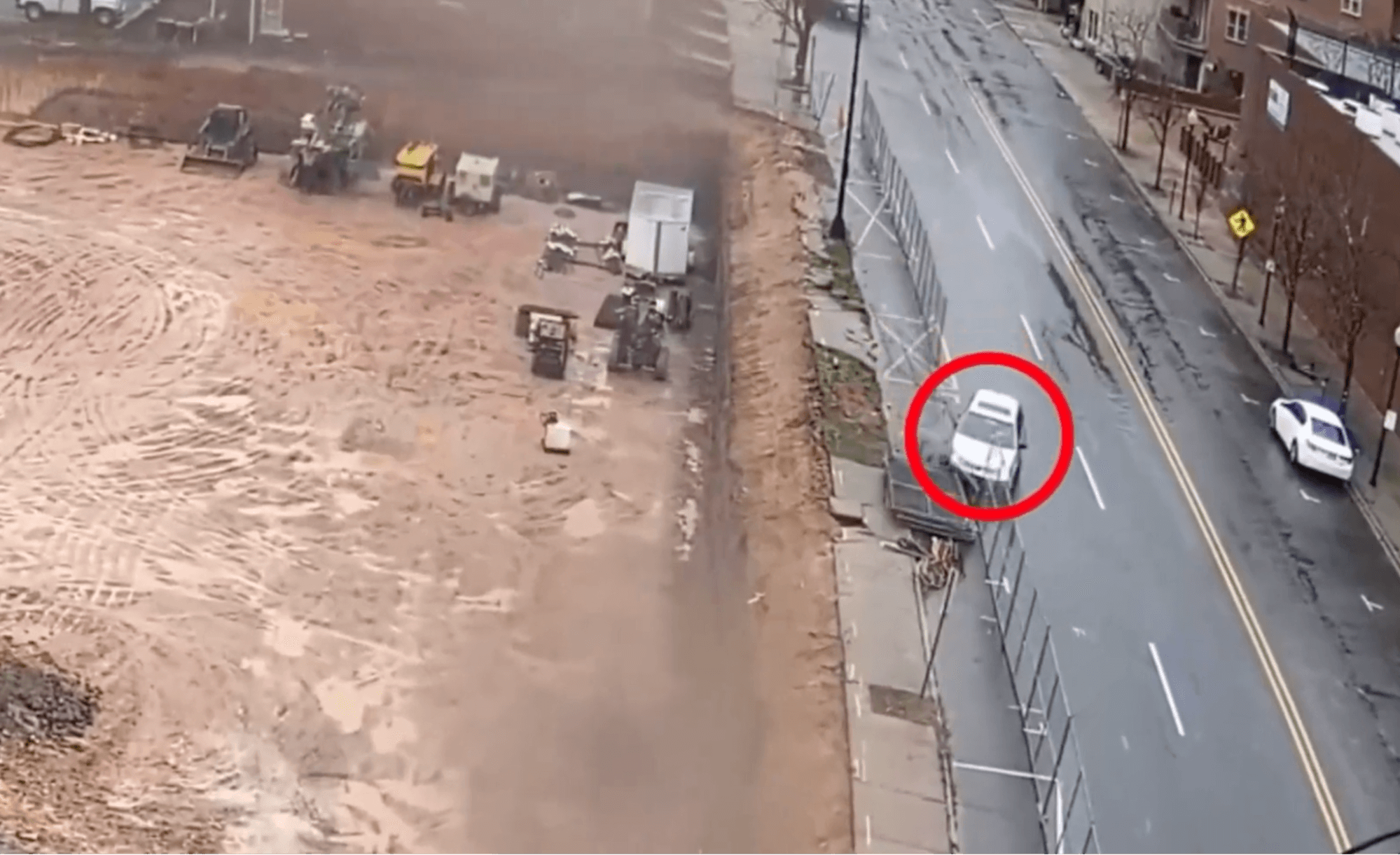 Aerial camera view showing a vehicle stopped near a fenced construction site.