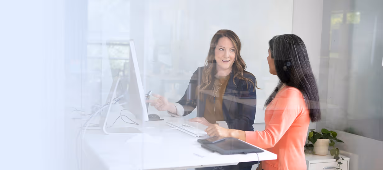 Zwei Frauen diskutieren, während sie an einem weißen Schreibtisch mit einem Computer in einem hellen Büro sitzen.