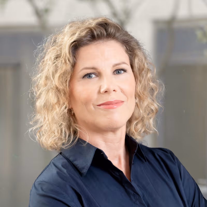 Coach Iris Kuenzi – A woman with curly blonde hair wearing a dark shirt, smiling slightly against a blurred background.