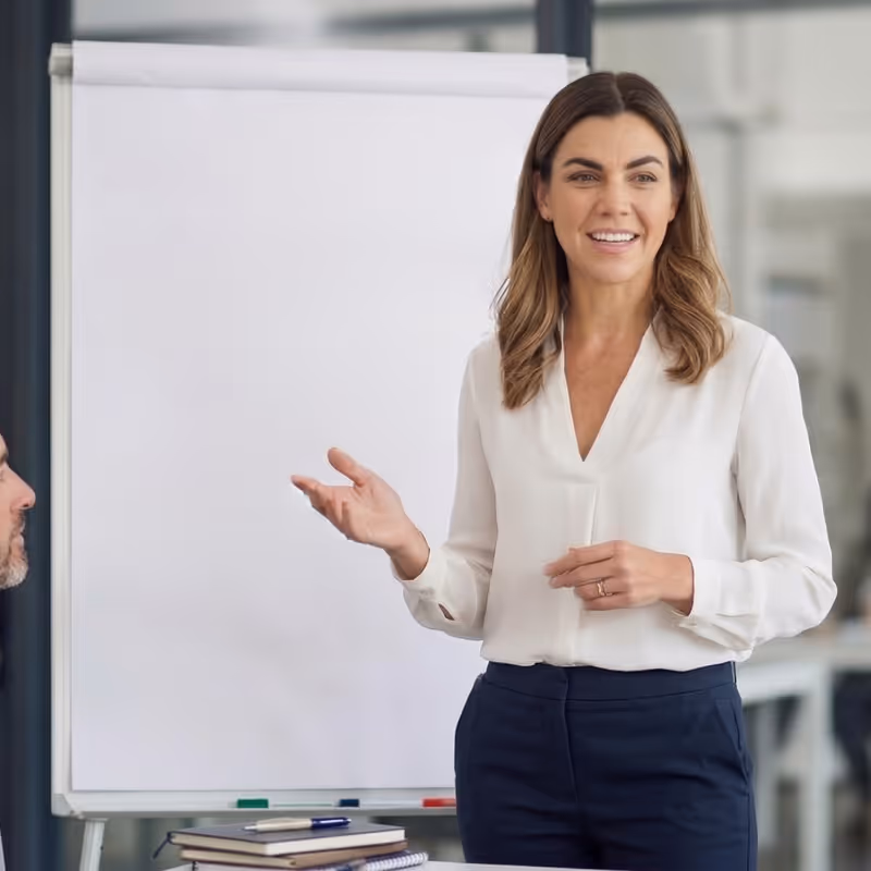 Woman in white blouse giving presentation near blank flip chart in office setting.