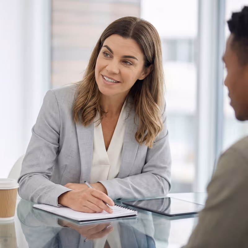 Smiling businesswoman in gray blazer taking notes while talking to a man in a bright office.
