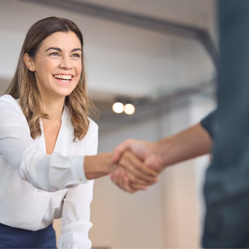 Smiling woman in white blouse shaking hands with another person in a modern office setting.