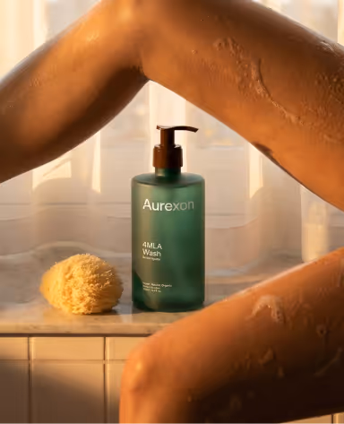 Bottle of Aurexon 4MLA Wash and a natural sponge by a window, framed by a person's bent arm and leg with visible dry skin.