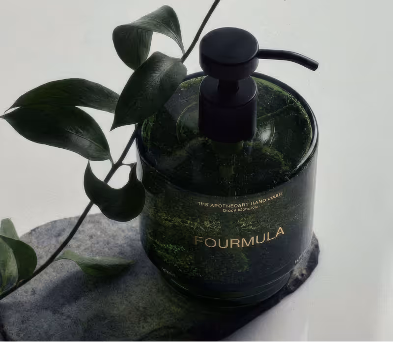 Dark green liquid soap dispenser bottle labeled 'FOURMULA' with a black pump and a leafy plant branch beside it.