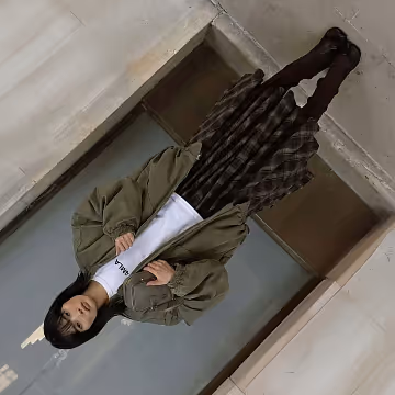 Person wearing an oversized green jacket, white t-shirt, plaid skirt, and black tights standing against an industrial-style concrete wall.