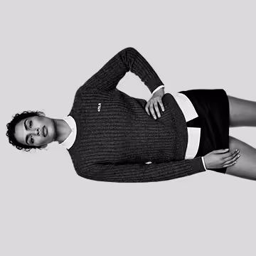 Black and white photo of a woman wearing a dark textured sweater and a short skirt, posing with one hand on her hip against a plain background.