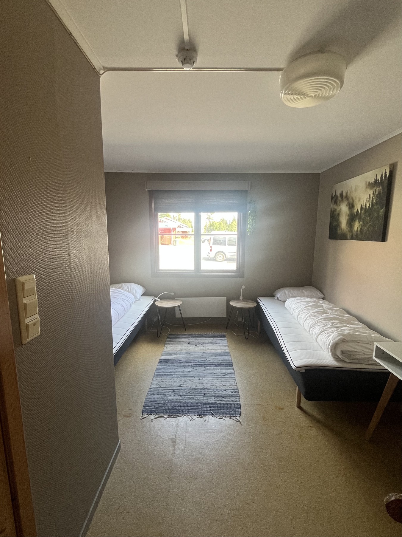 Lite hotellrom med to enkeltsenger, nattbord og et vindu med utsikt til parkeringsplassen.
