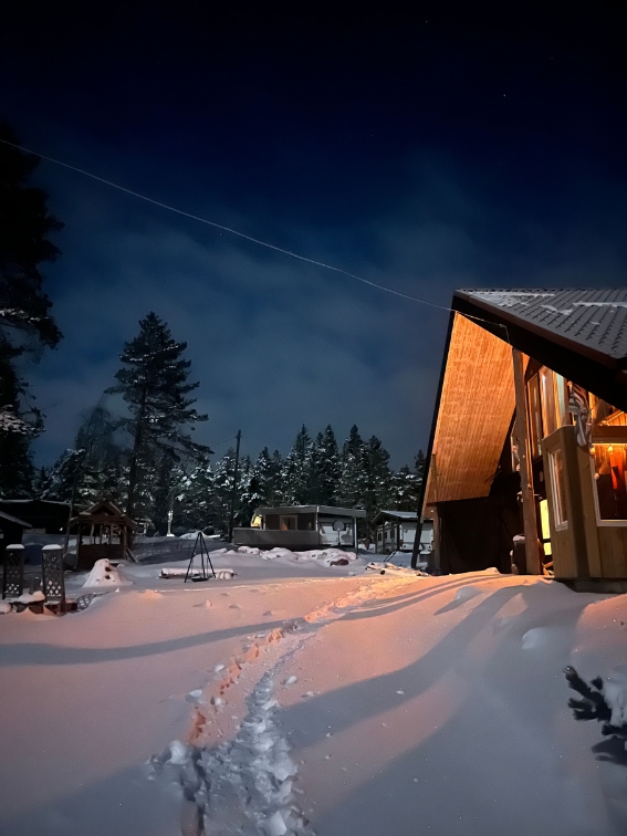 Snødekket hytte med opplyst veranda om natten og fotspor i snøen som leder mot et skogkledd område.