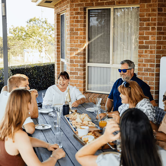 Group of six people sitting around an outdoor wooden table with plates and food, enjoying a casual gathering on a patio with a brick wall background.