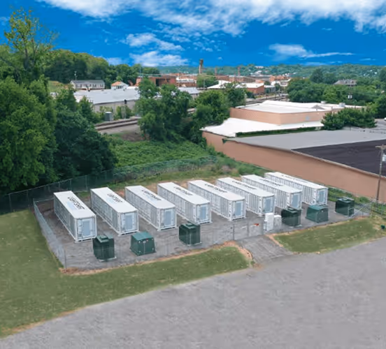 A fenced area containing eight white electrical or utility boxes arranged in two rows near industrial buildings and trees under a partly cloudy sky.