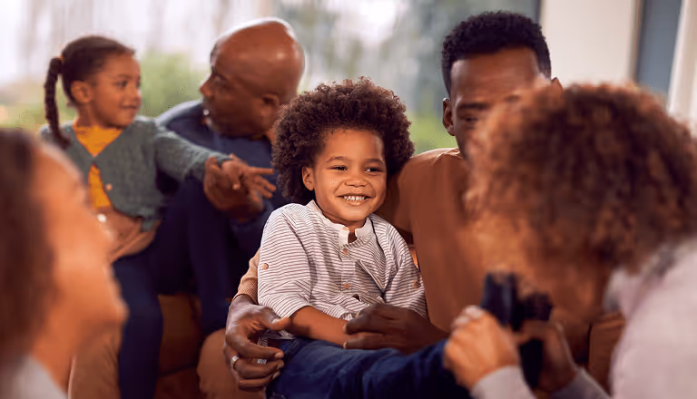 Smiling young boy sitting on a man's lap surrounded by family members interacting warmly.
