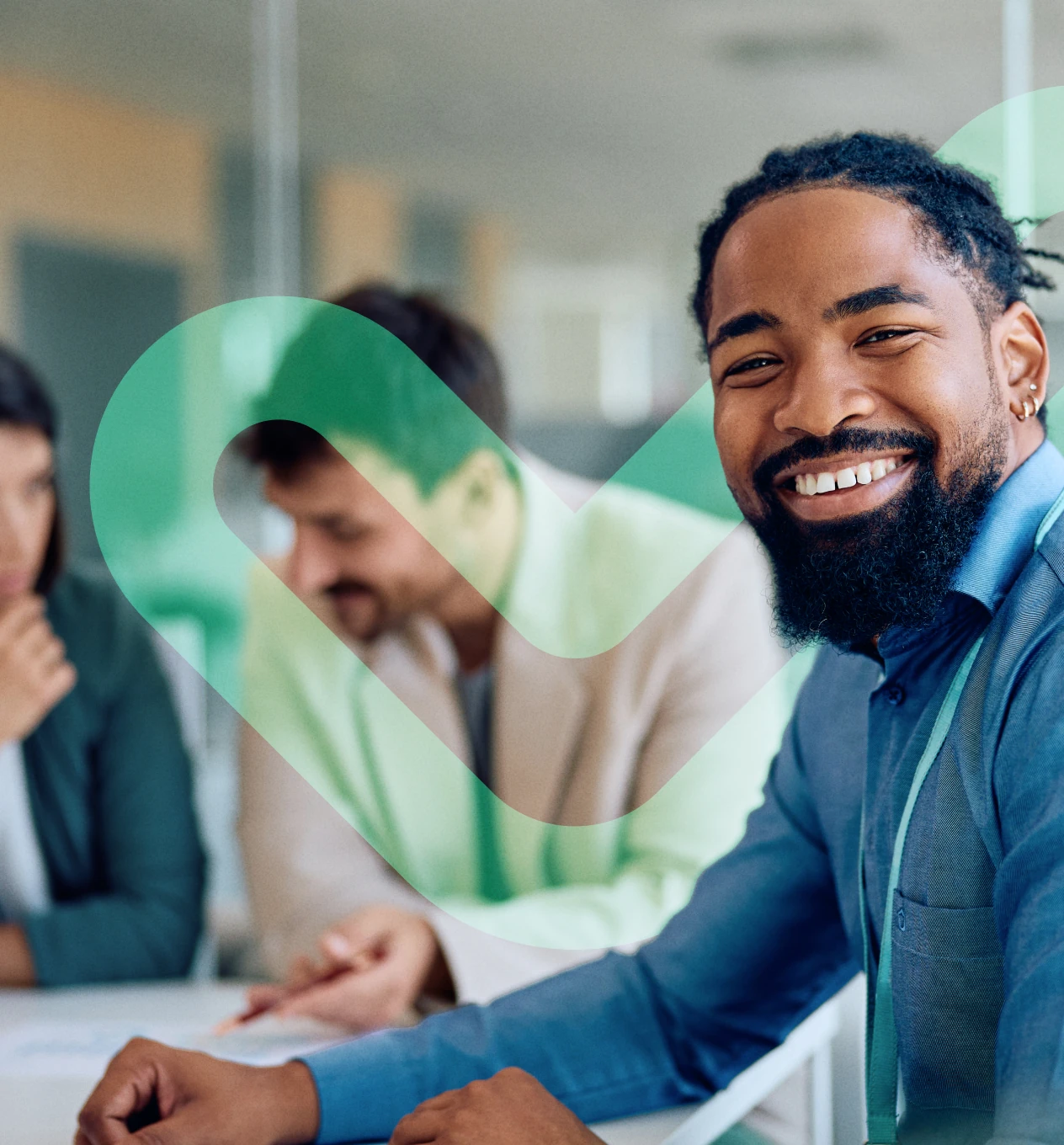 Smiling man with beard and blue shirt sitting at a table with two colleagues in a blurred office background.