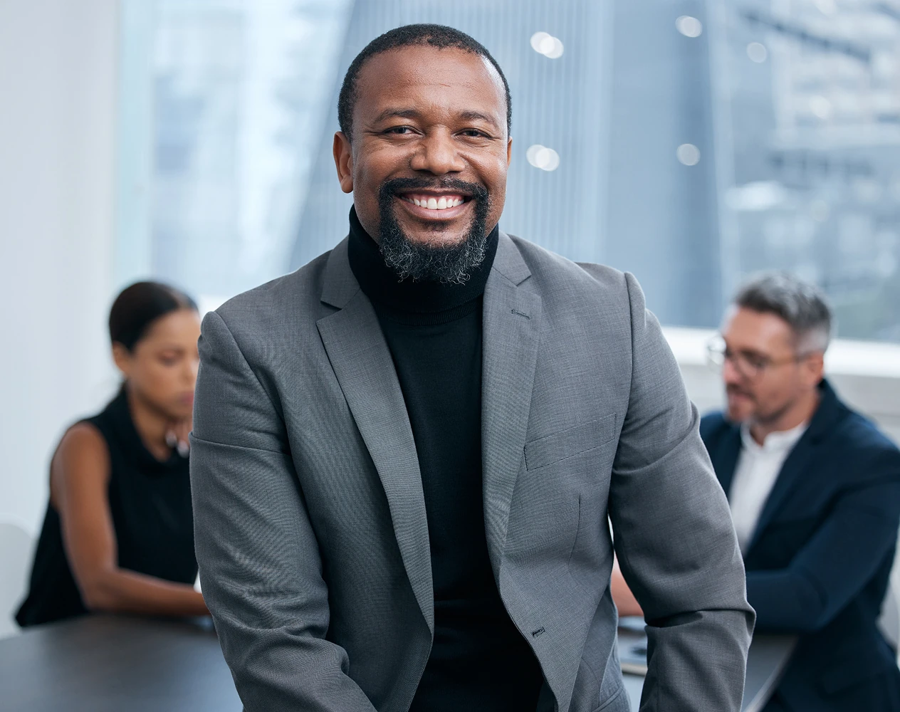 Smiling man in gray suit and black turtleneck sitting in front of a desk with two focused colleagues in a modern office.