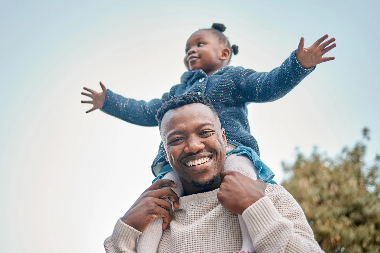 Smiling man carrying a child on his shoulders with the child spreading arms outdoors.