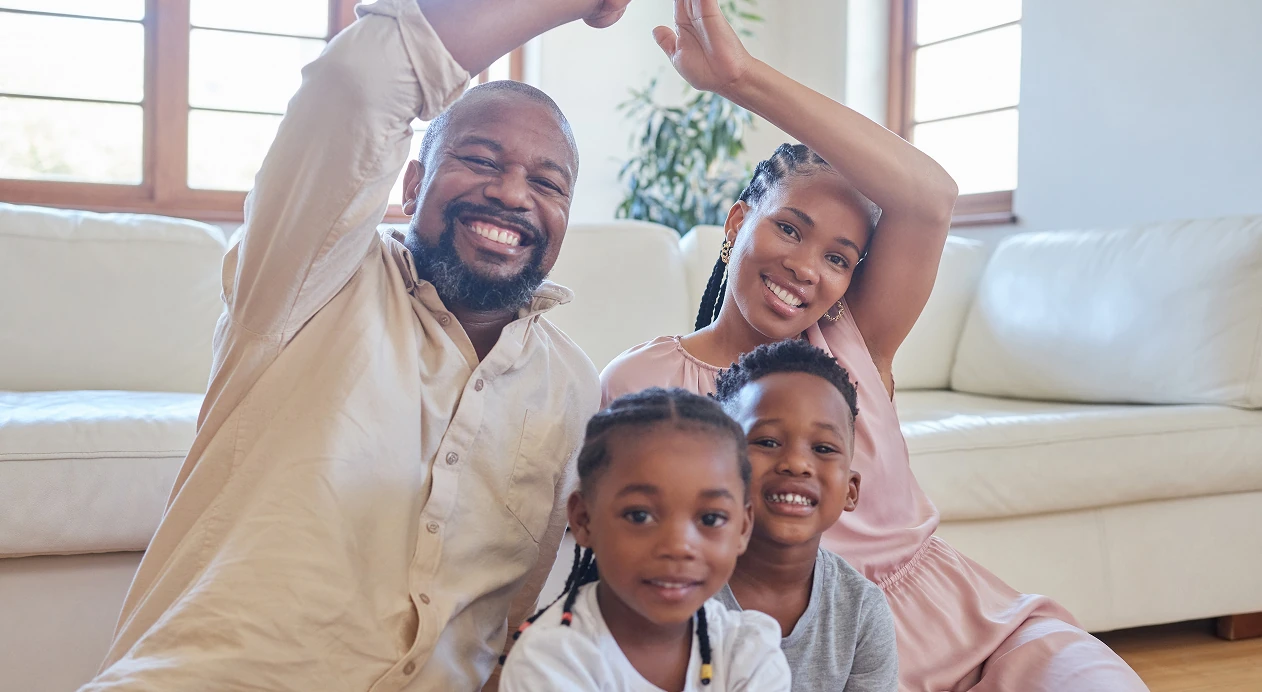 Smiling family of four sitting on the floor in a bright living room, raising their arms cheerfully.
