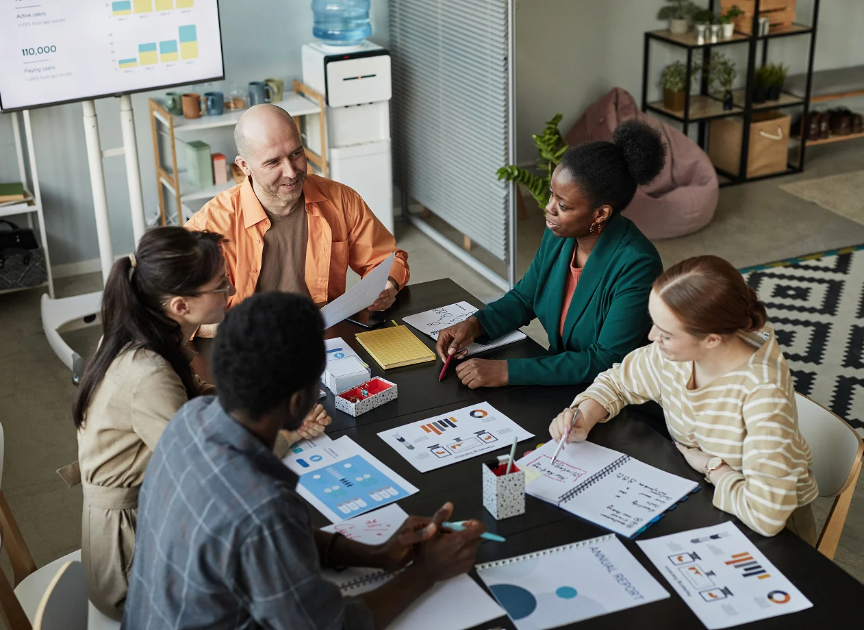 Five diverse colleagues sitting around a table reviewing charts and notes during a meeting in a modern office.