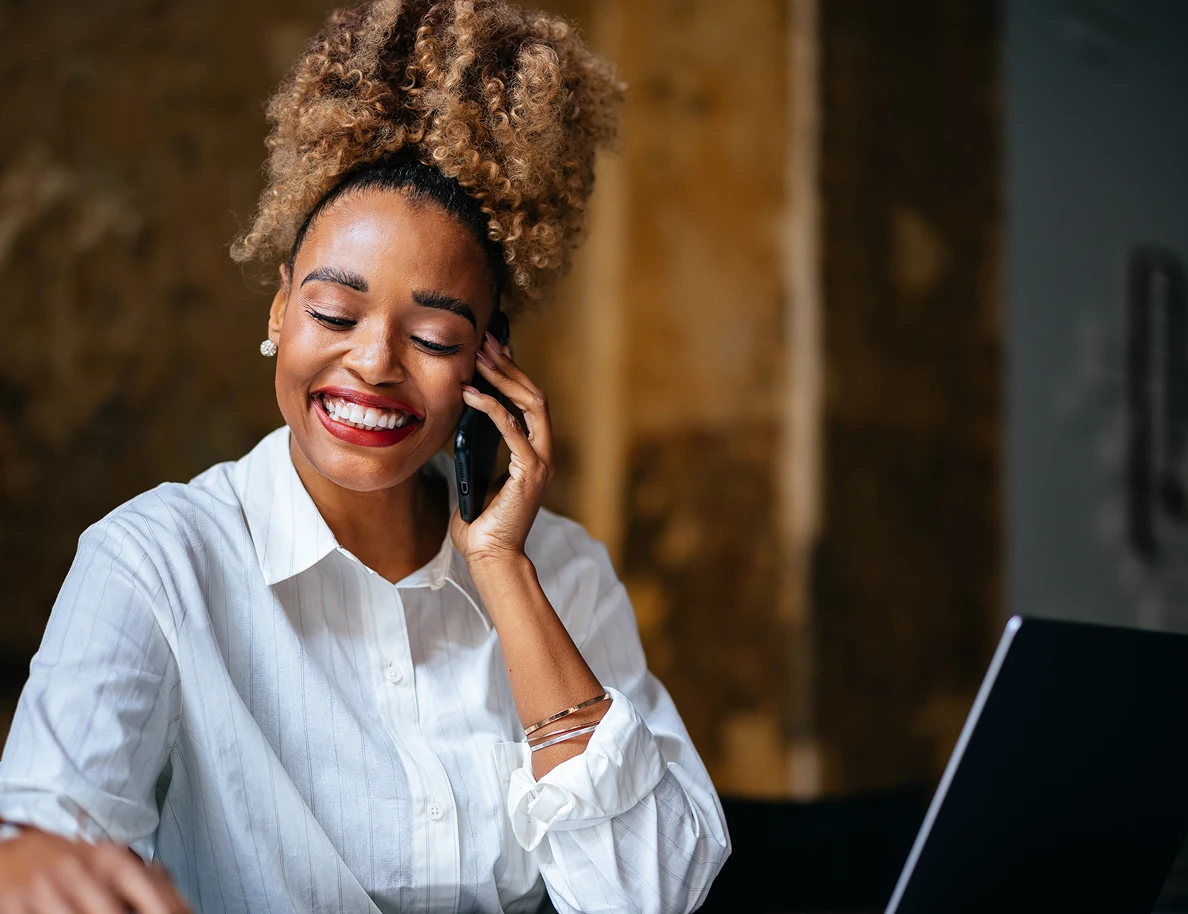 Smiling woman with curly hair in a white shirt talking on a smartphone, sitting at a desk with a laptop.