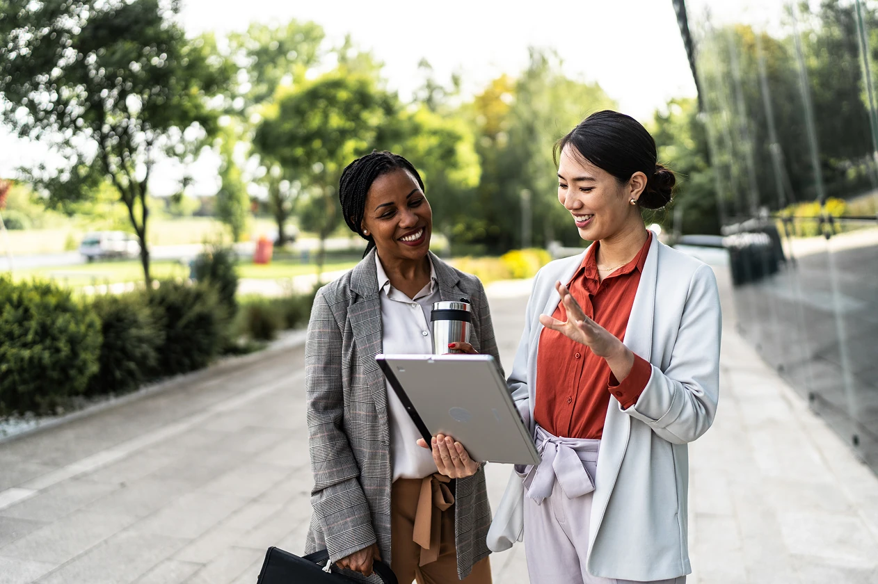 Two professional women standing outdoors, looking at a laptop and smiling, one holding a travel mug.