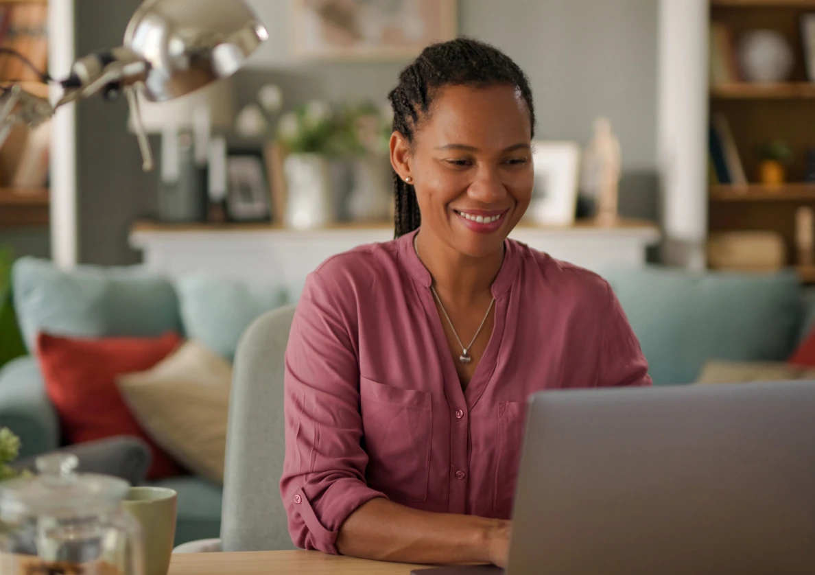 Smiling woman in a pink shirt working on a laptop at a wooden table in a cozy living room.