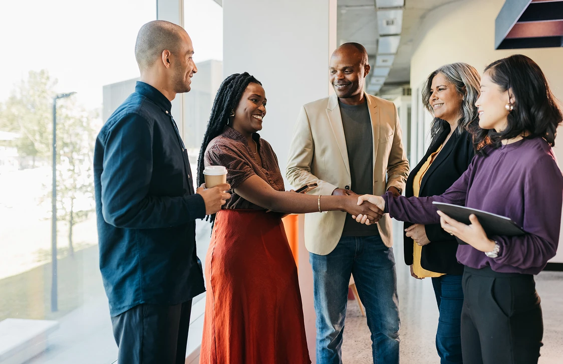 Five diverse professionals smiling and shaking hands in a modern office hallway with large windows.