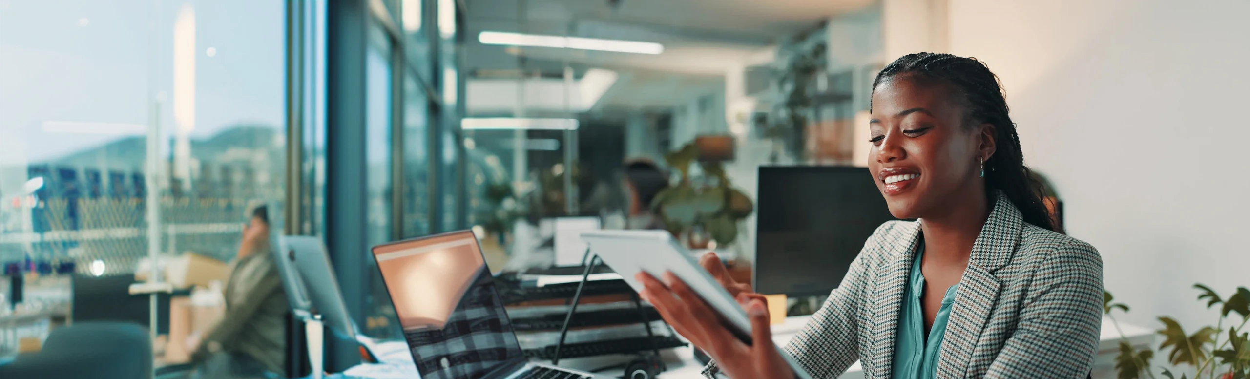 Smiling woman using a tablet in a modern office with laptops and large windows.