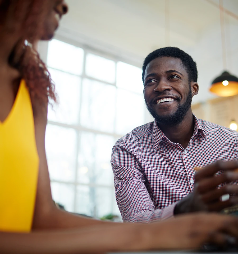 Smiling man in a checked shirt conversing with a woman in a yellow top near a large window.