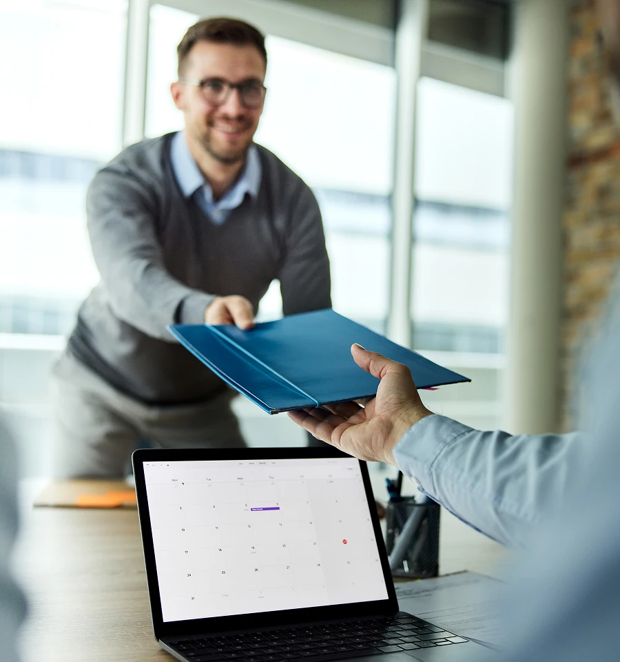 Man in glasses handing a blue folder to another person across a table with an open laptop showing a calendar.