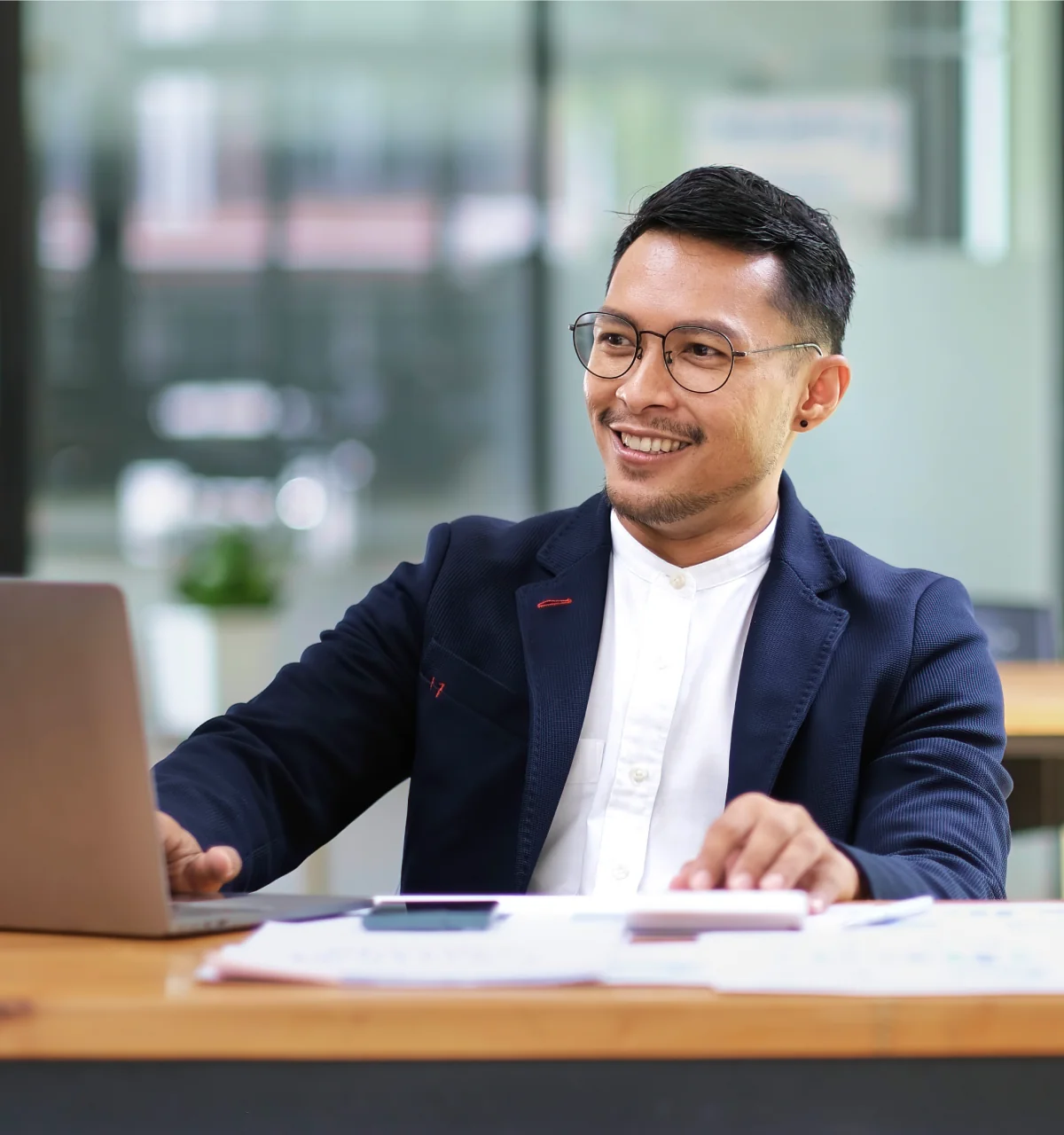 Smiling man wearing glasses and a navy blazer working on a laptop at a desk.