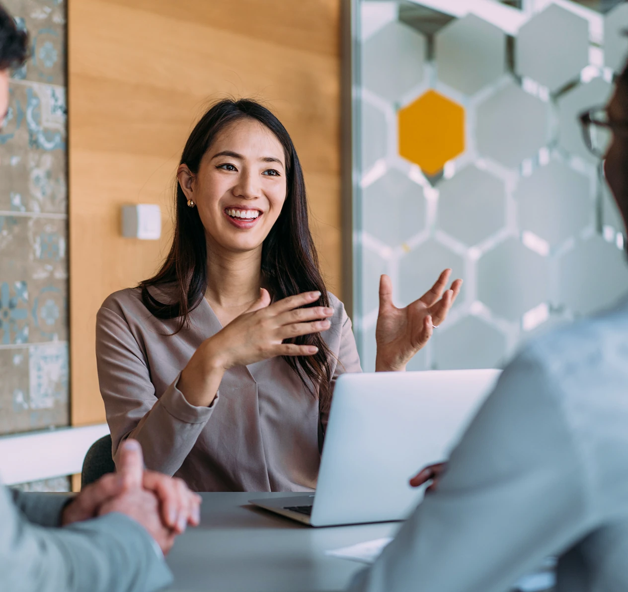 Smiling woman gesturing with hands while talking to two people across a table with a laptop.