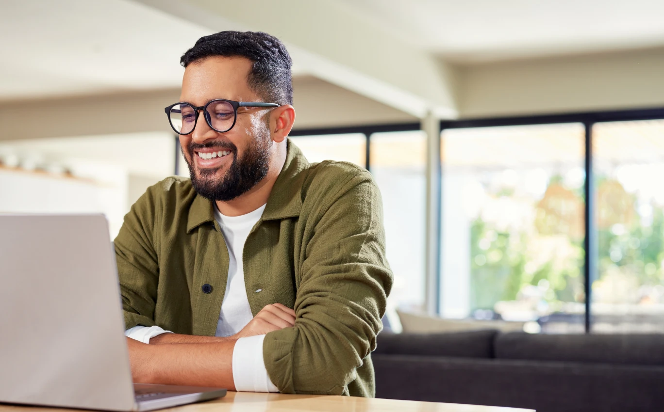 Smiling bearded man with glasses using a laptop at a wooden table in a bright living room.