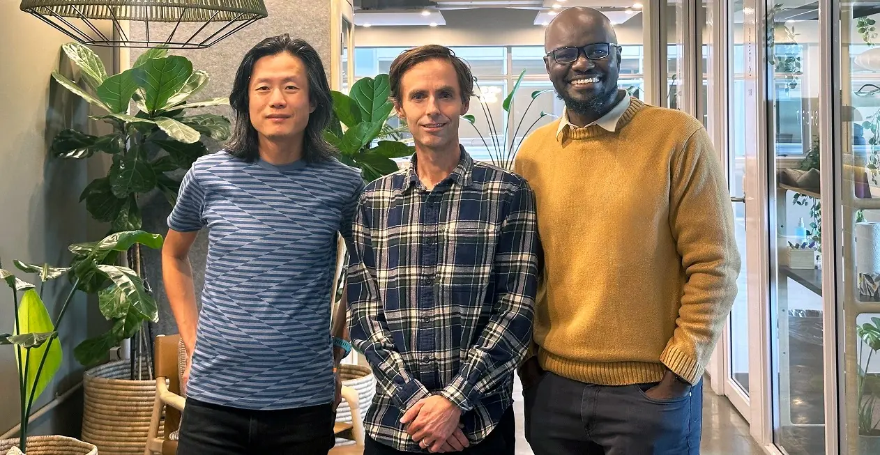 Three men standing side by side indoors, smiling, with plants and office furniture in the background.