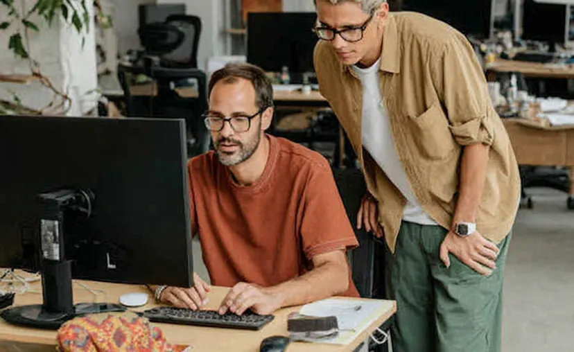 Two men wearing glasses working together at a computer in an office setting.