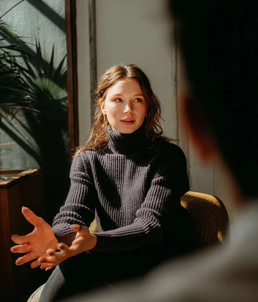 Young woman in a black turtleneck sweater gesturing while talking in a sunlit room.