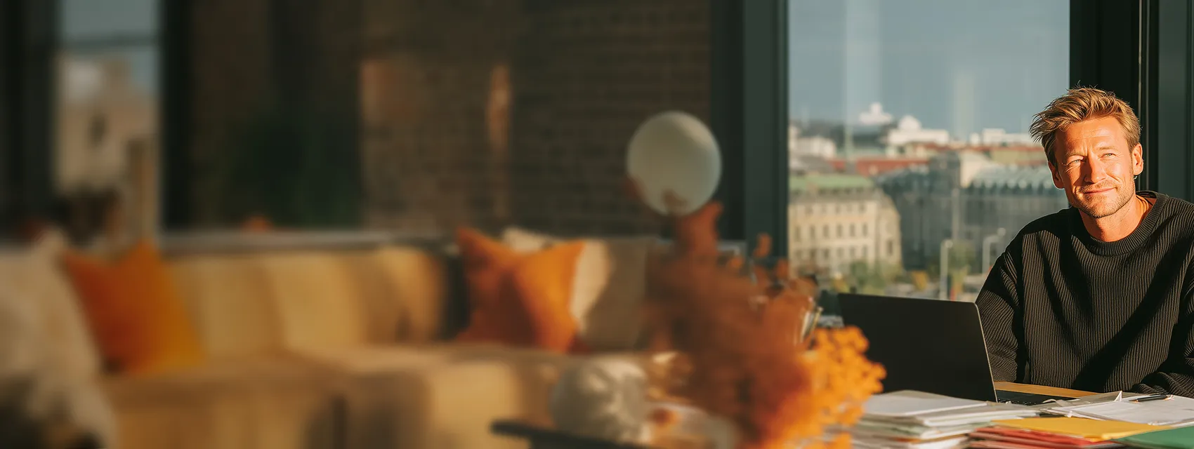 Man smiling while sitting at a desk with a laptop and paperwork in a sunlit room with a window view of buildings.