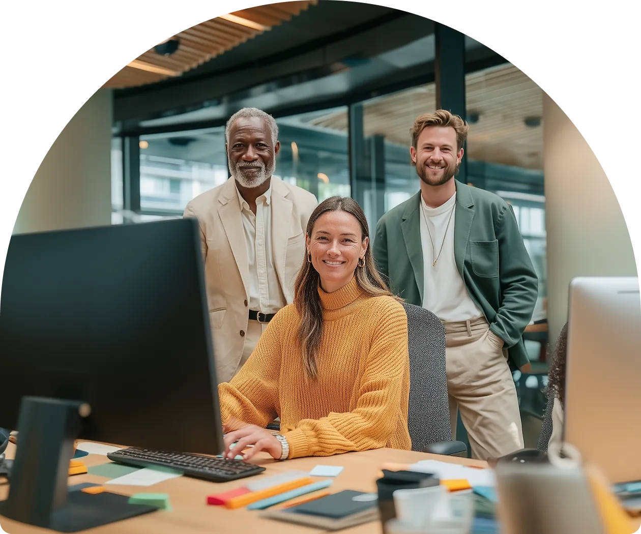 Three colleagues smiling in a modern office with a woman in a yellow sweater typing at a computer and two men standing behind her.