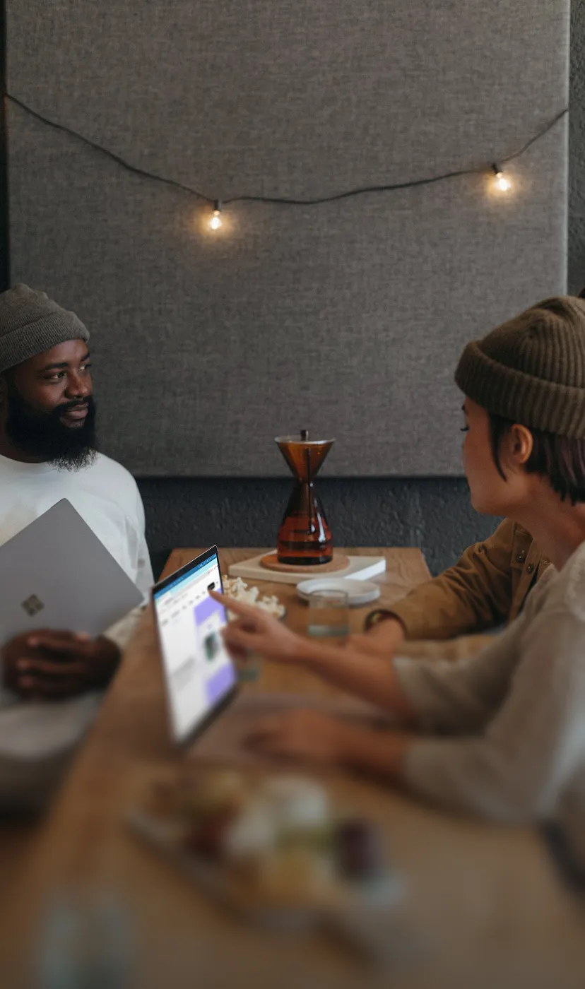 Two people sitting at a wooden table with a laptop, one pointing at the screen while the other holds a tablet, with string lights and a coffee carafe in the background.