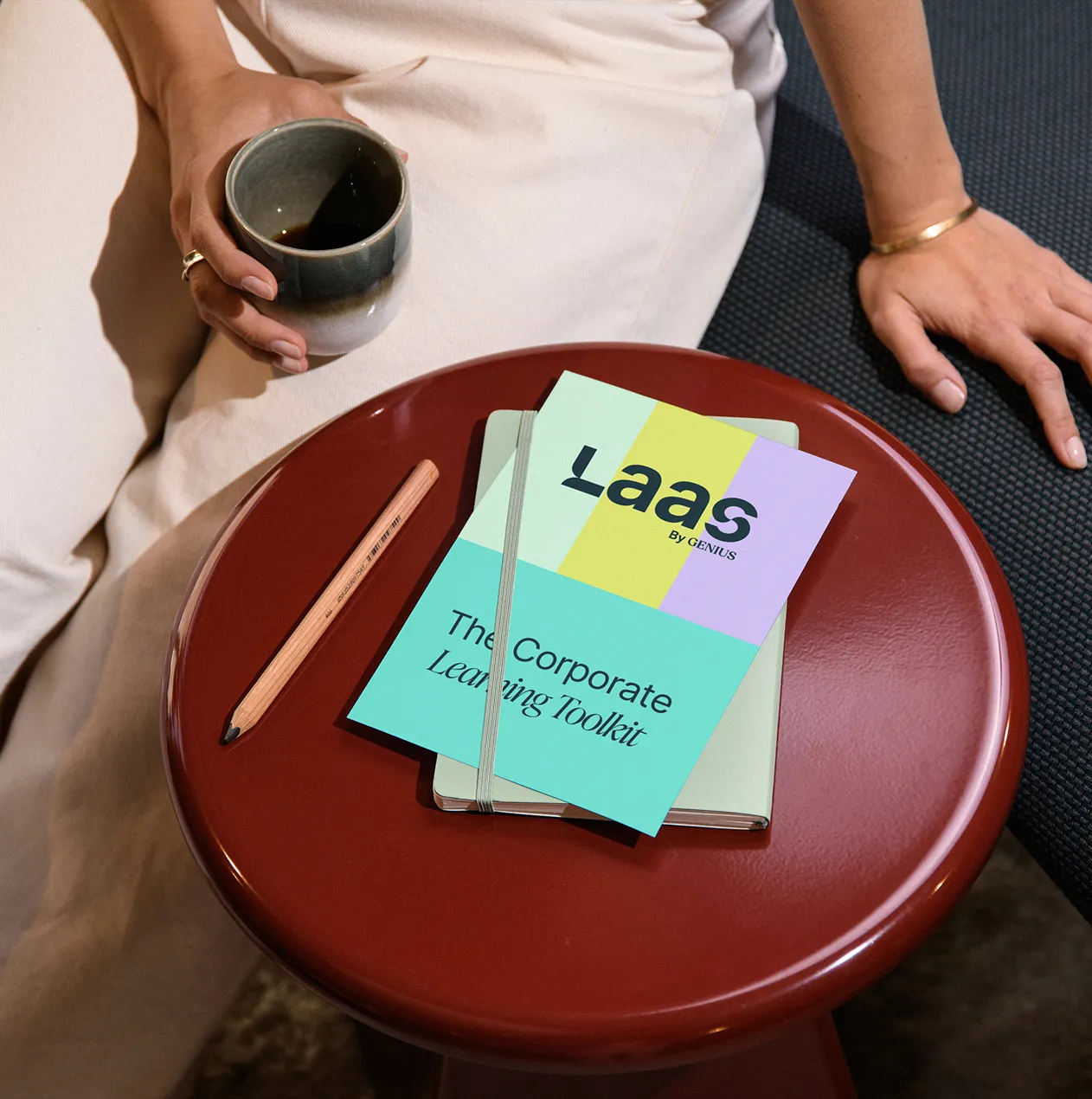 Person holding a cup of black coffee sitting next to a red round table with a wooden pencil and a notebook labeled 'Laas by Genius: The Corporate Learning Toolkit'.