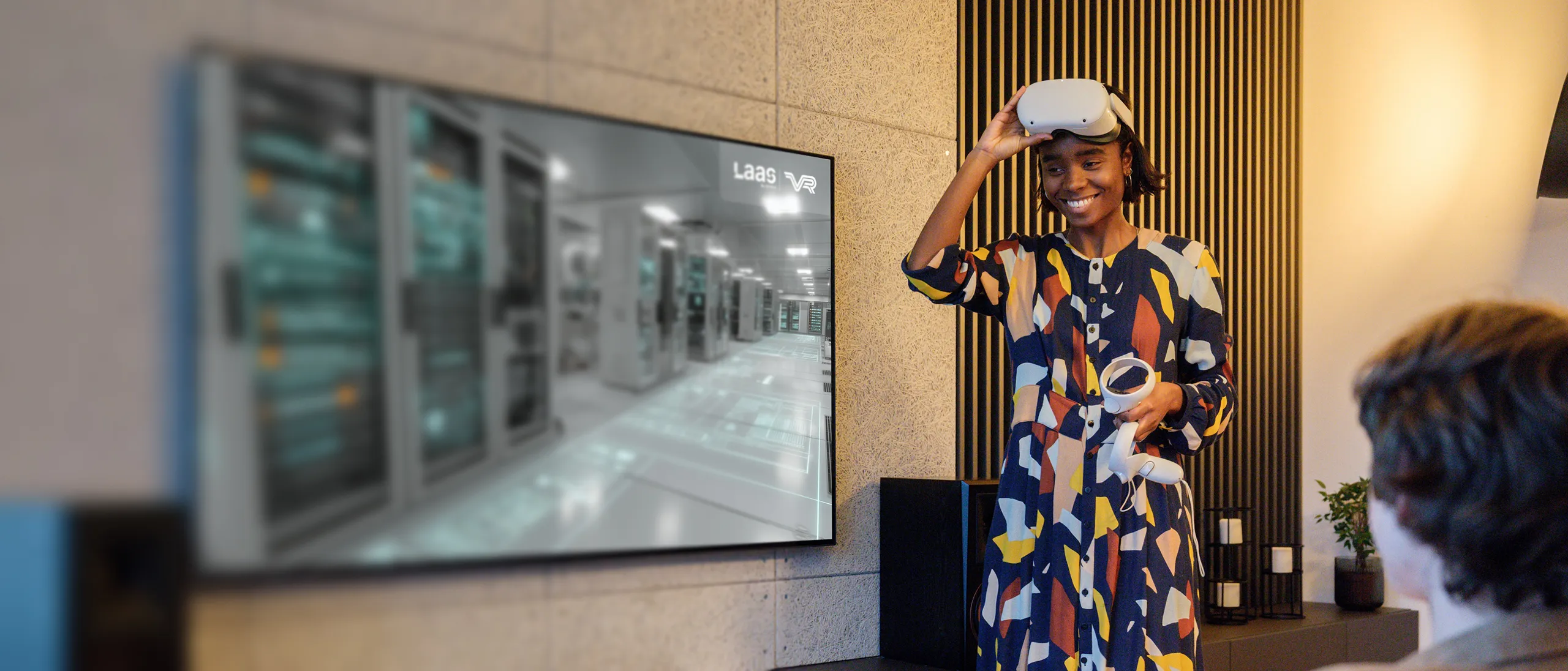 Smiling woman holding VR controllers and lifting a VR headset from her head in a modern room with a large screen displaying a data center image.